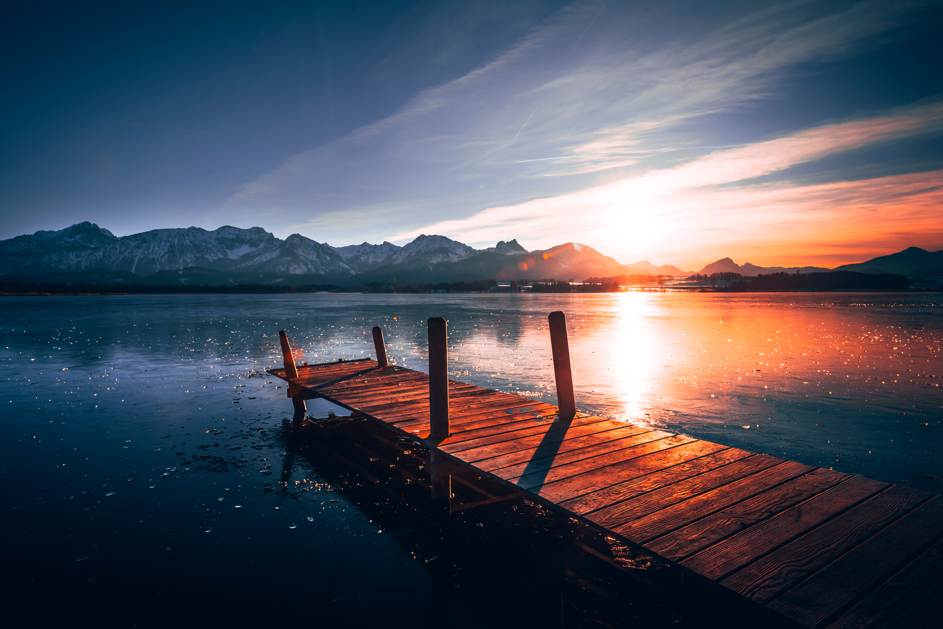 Holzsteg über dem gefrorenen, winterlichen Hopfensee im Ostallgäu bei einem farbenprächtigen Sonnenuntergang über den Allgäuer Alpen